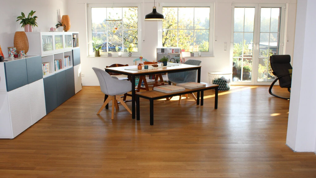 Aufnahme in ein Wohnzimmer mit einem Esstisch in der Mitte, einem Sideboard auf der linken Seite und einem Stuhl auf der rechten Seite. Beim Boden handelt es sich um einen braunen Holzboden und im Hintergrund sind große Fenster zu erkennen.