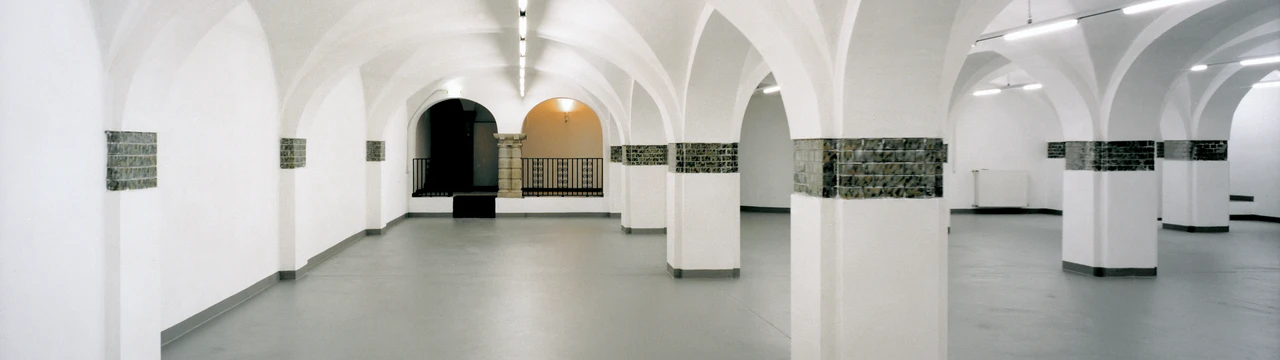 Weitläufiges Kellergewölbe mit Säulen, weißen Wänden und grauem Boden