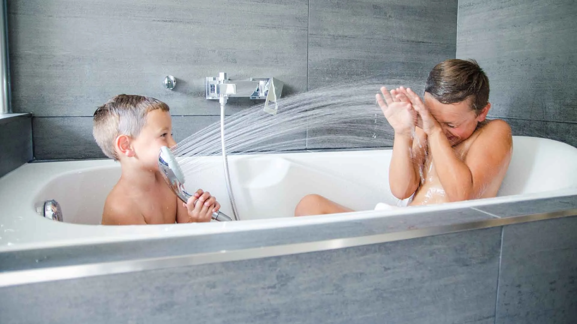 Zwei Jungen spielen in einer grau gefliesten Badewanne mit dem Duschkopf