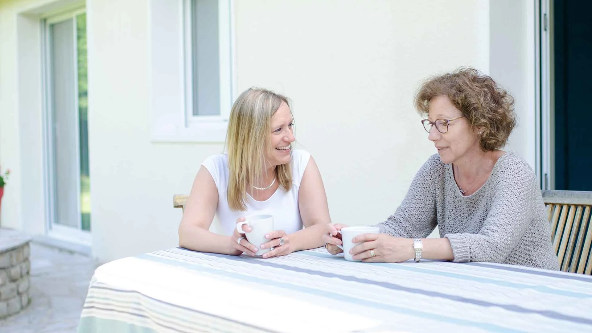 Zwei Frauen sitzen an einem Gartentisch auf der Terrasse vor einer hellgelben Fassade