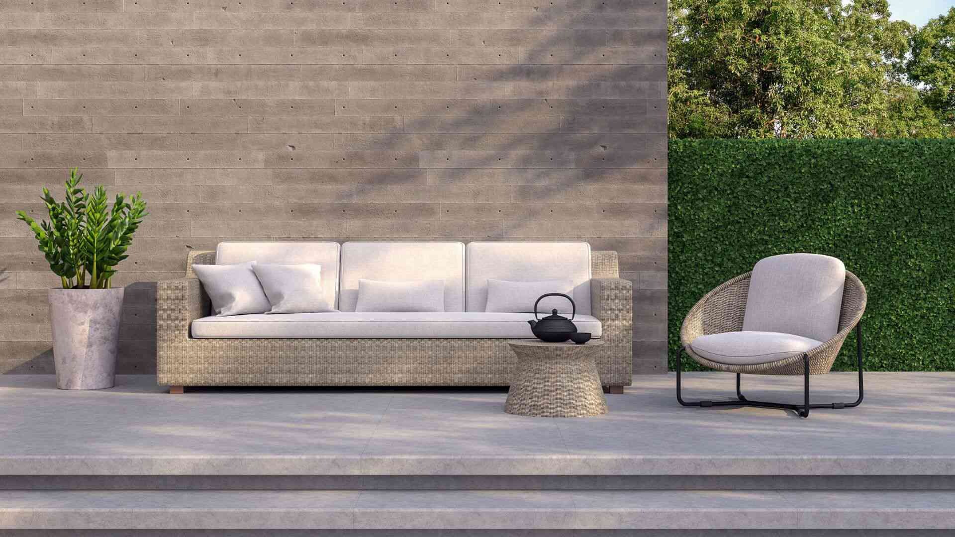 Terrasse Gestaltung einer modernen Terrasse mit Fliesenboden, einem Sofa mit hellen Kissen und einer Topfpflanze vor einer Hauswand, an die eine Hecke anschließt