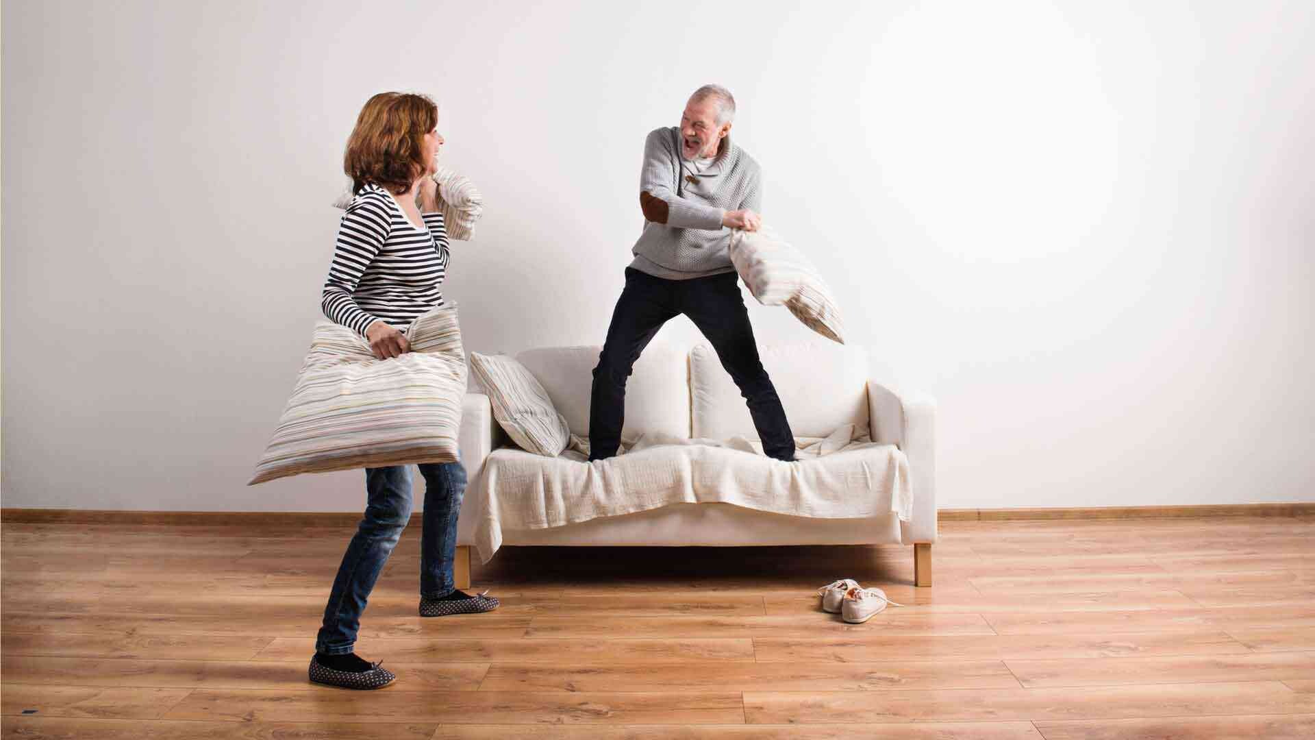 Trittschalldämmung Mann und Frau machen eine Kissenschlacht auf einem Sofa vor einer weißen Wand auf Holzfußboden