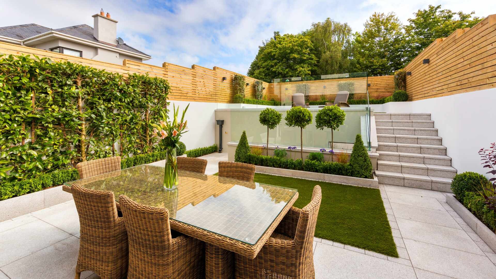 Garten mit gefliester Terrasse Blick in einen mehrstufigen Garten mit Holzzaun, grünen Beeten, gefliester Terrasse und Korbmöbeln