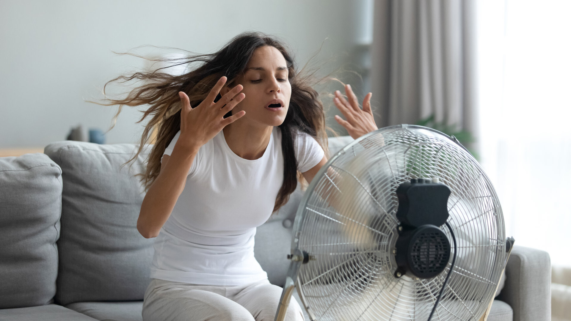 Sommerlicher Wärmeschutz Junge Frau sitzt auf einem Sofa vor einem Ventilator und lässt sich den Wind ins Gesicht pusten