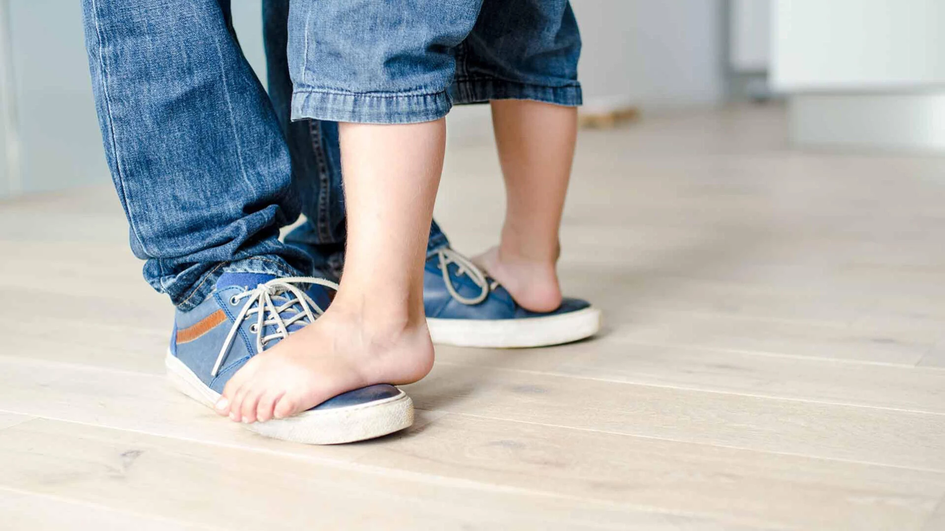 Bild von einem Holzfußboden auf dem ein kleineres Paar nackter Füße auf einem größeren Paar Füße mit Schuhen steht