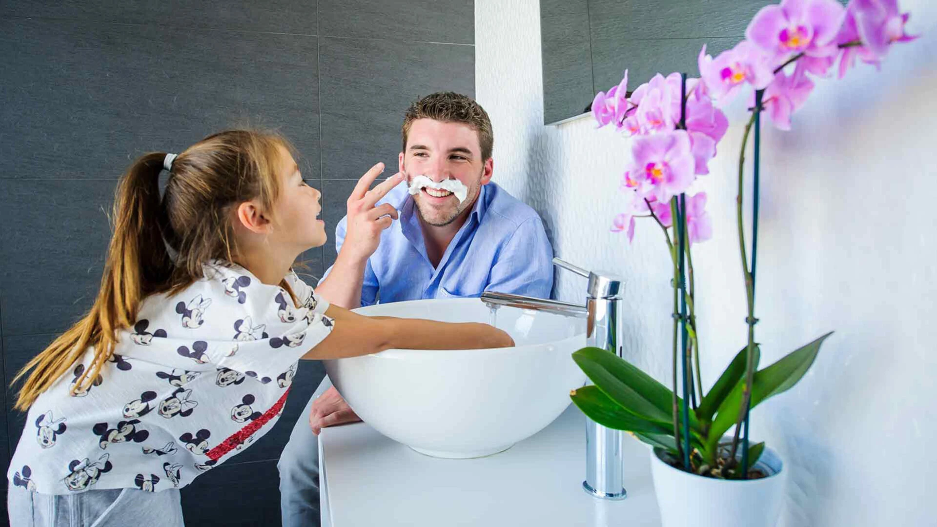 Vater und Tochter sind am Waschbecken im Badezimmer und der Vater hat einen Bart aus Rasierschaum im Gesicht