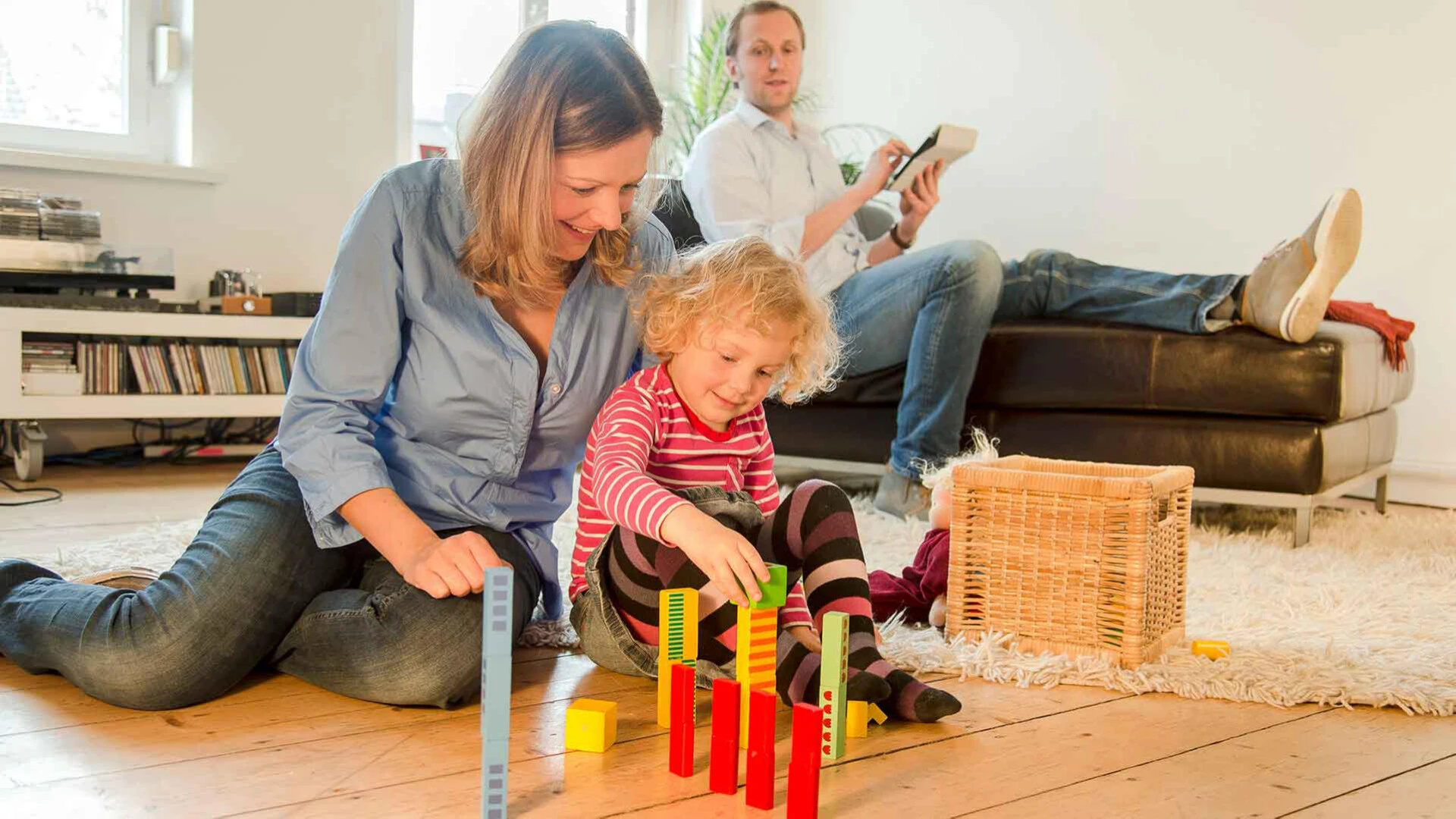 Aufnahme eines Wohnzimmers, in dem die Mutter mit ihrem Kind auf dem Boden spielt und der Vater auf dem Sofa am Tablet ist