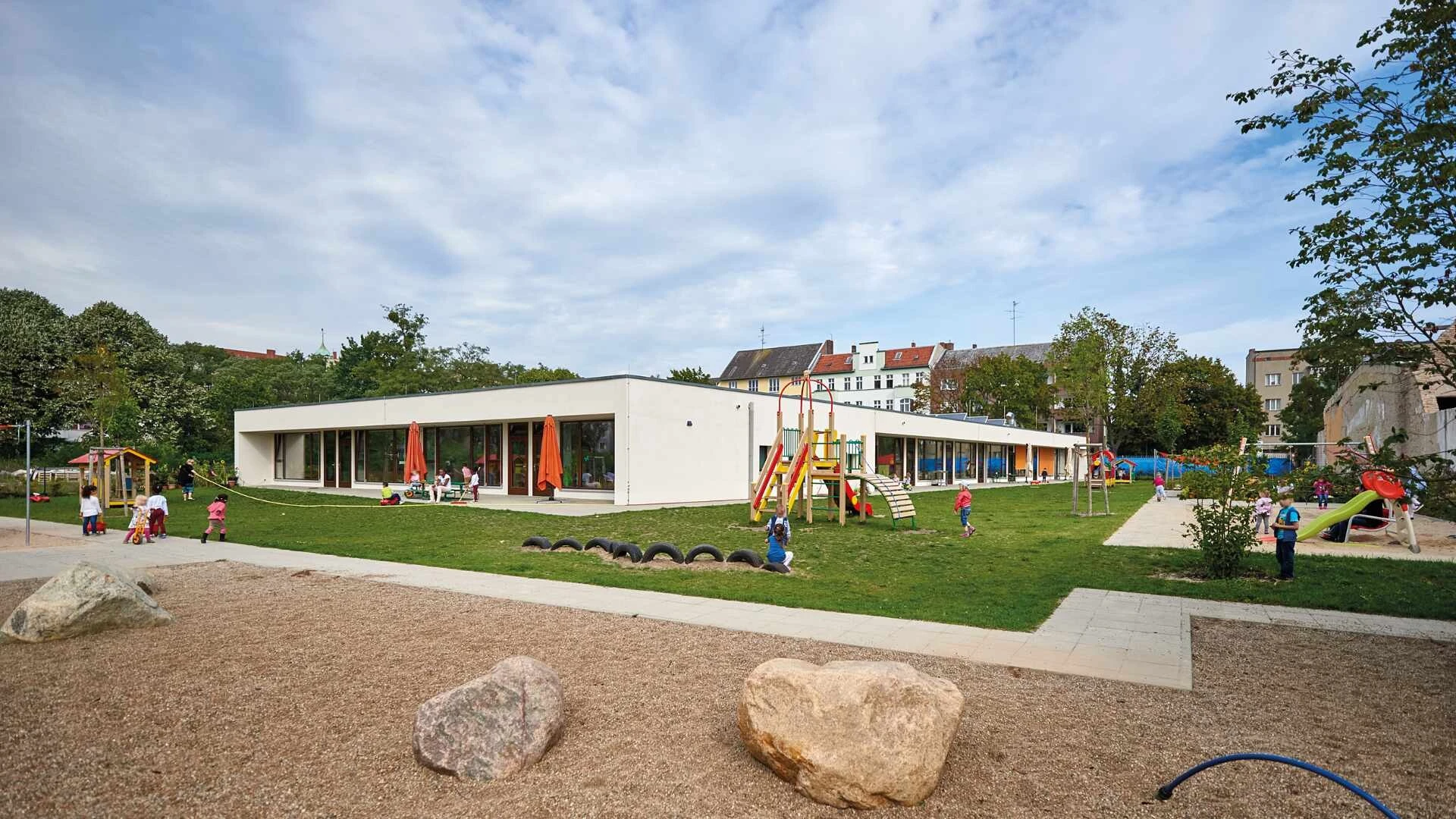 Weitwinkelaufnahme eines Kita-Gebäudes mit spielenden Kindern drum herum