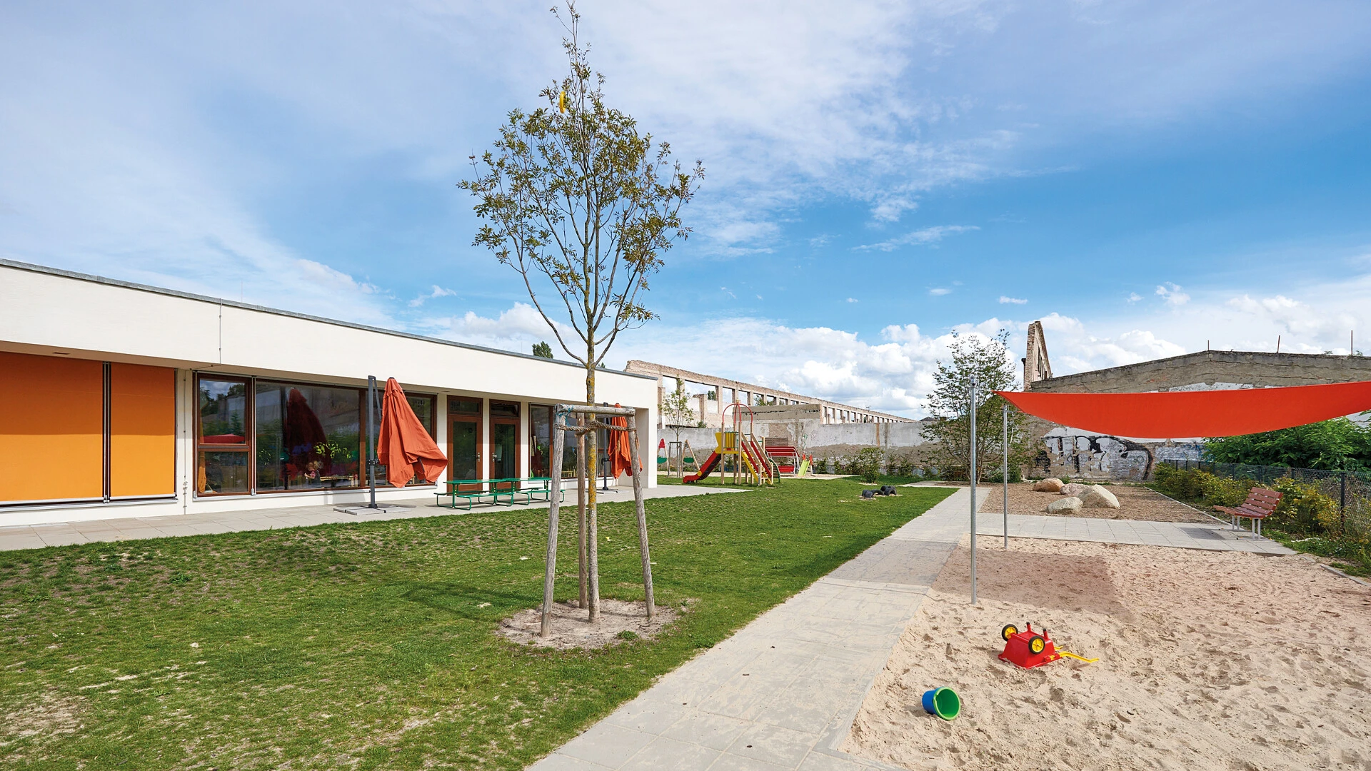 Außenansicht auch eine Kita mit gedämmter Fassade, Raffstoren an den Fenstern und Rasenfläche und beschattetem Sandkasten