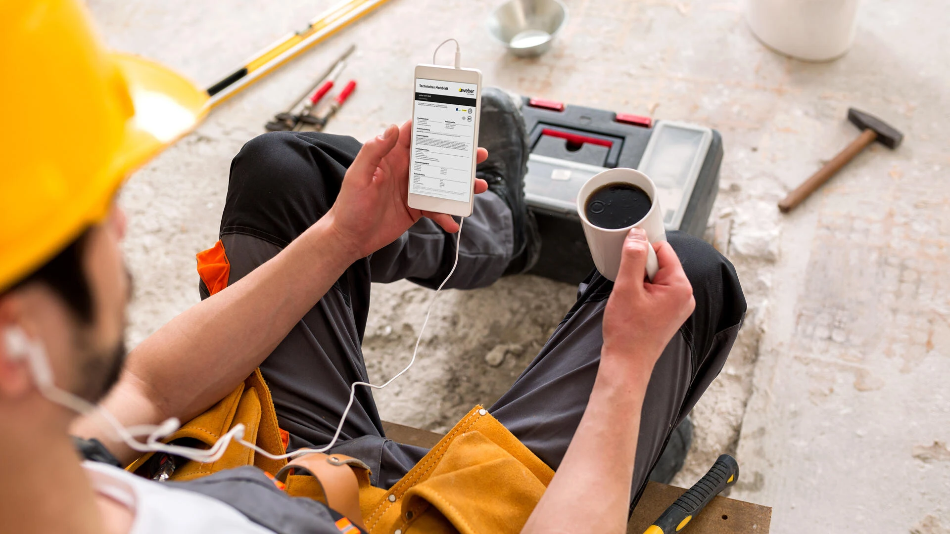 Bauarbeiter mit gelbem Helm sitzt auf der Baustelle mit Kaffee und Handy in der Hand, wo der Weber Messenger geöffnet ist
