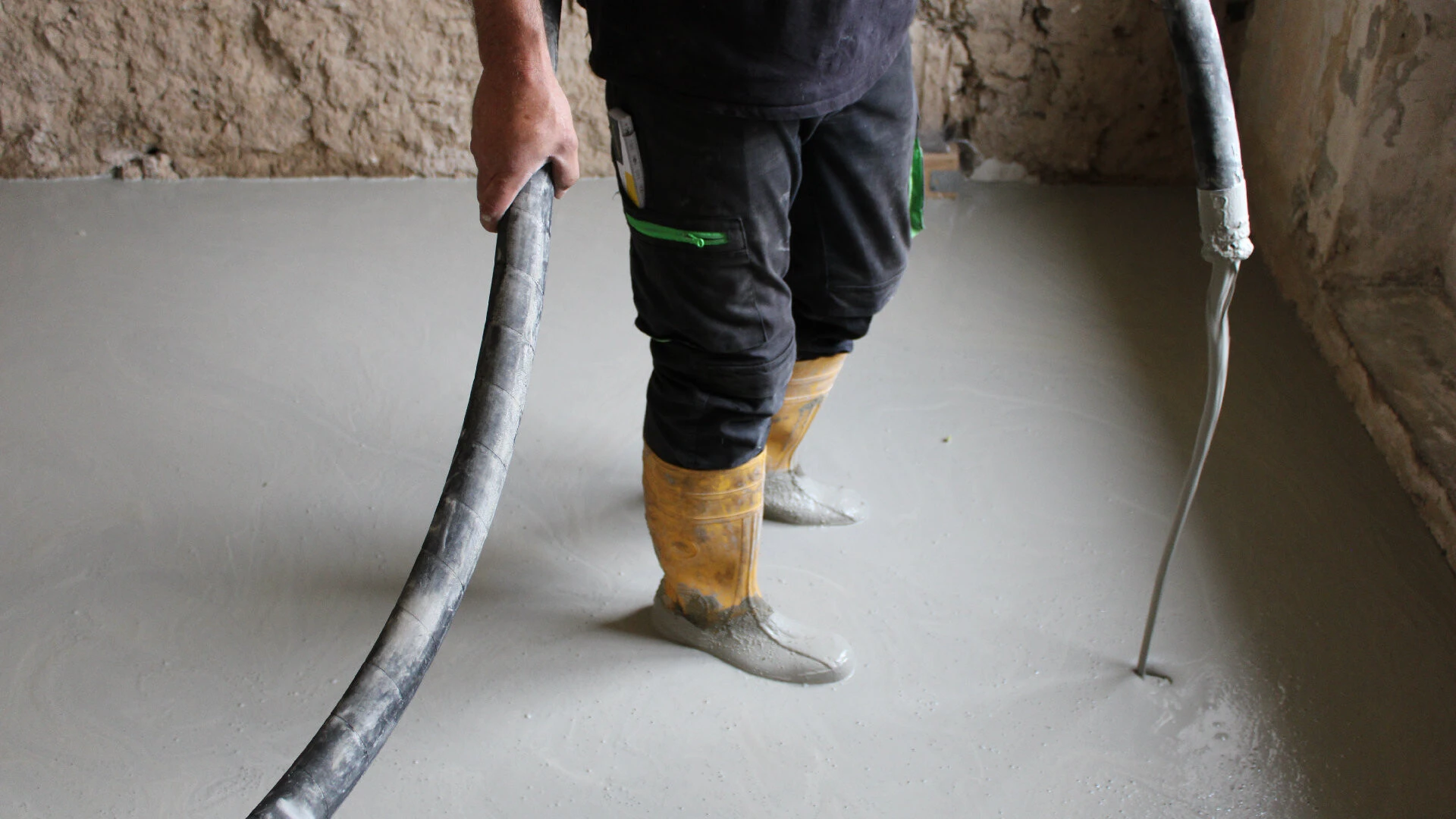 Im Bild sieht man zwei Beine in Arbeitskleidung und gelben Gummistiefeln, die in einer frischen Fläche des Höhenausgleichs weber.floor 4305 stehen. Außerdem sieht man einen Schlauch, der von links unten in die Mitte oben läuft, in der Mitte oben von einer Hand gehalten wird und dann auf der rechten Seite von den Beinen wieder im Bild ist. Auf der rechten Seite von den Beinen ist das Ende des Schlauchs, aus dem frischer Höhenausgleich läuft