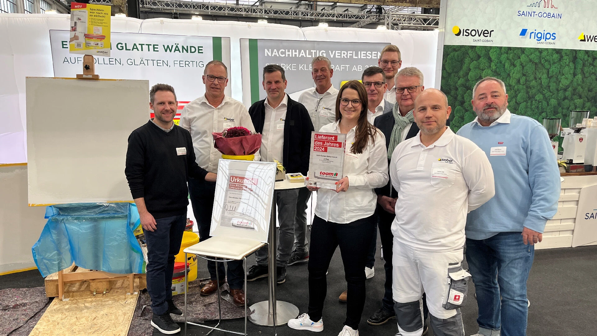 Aufnahme einer Gruppe von Mitarbeitenden von Saint-Gobain Weber aus dem DIY Bereich, die auf einem Messestand stehen. In der Mitte der Gruppe steht eine junge Frau mit einer Auszeichnung in der Hand