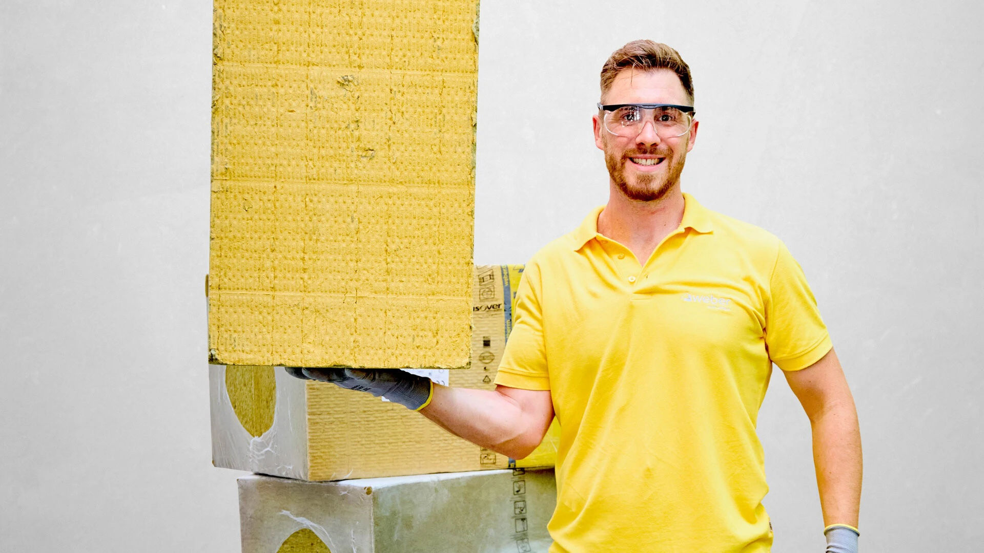 junger Mann steht frontal zur Kamera, er trägt ein gelbes Poloshirt, eine Schutzbrille und Handschuhe, in seiner  rechten Hand hält er einen Stapel Dämmplatten, er dteht vor einer weißen Wand, im Hintergrund sind weitere Stapel der Platten