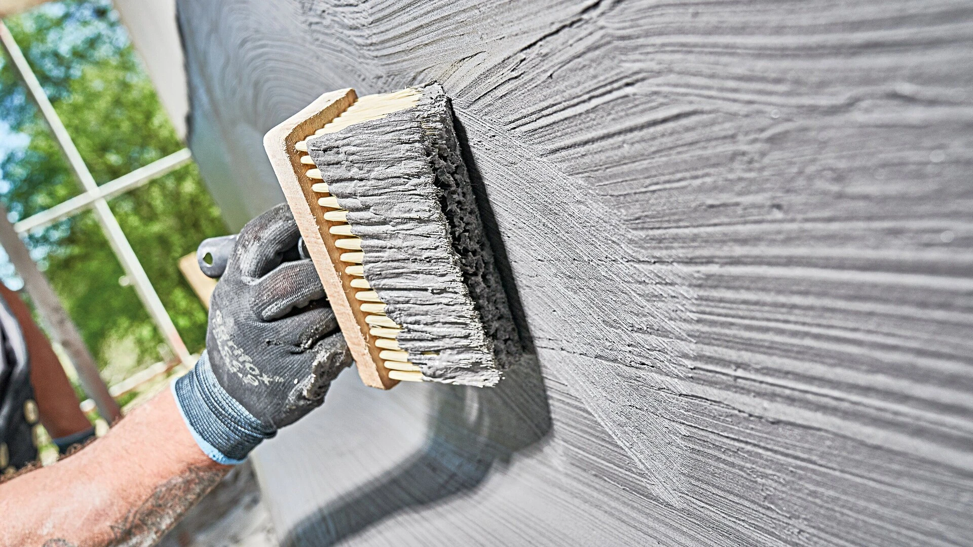 Nahaufnahme einer Hand mit Arbeitshandschuh, die eine Bürste über eine frisch verputzte Wand führt, um die Oberfläche zu strukturieren. Die Wand zeigt gleichmäßige Rillen im grauen Putz.