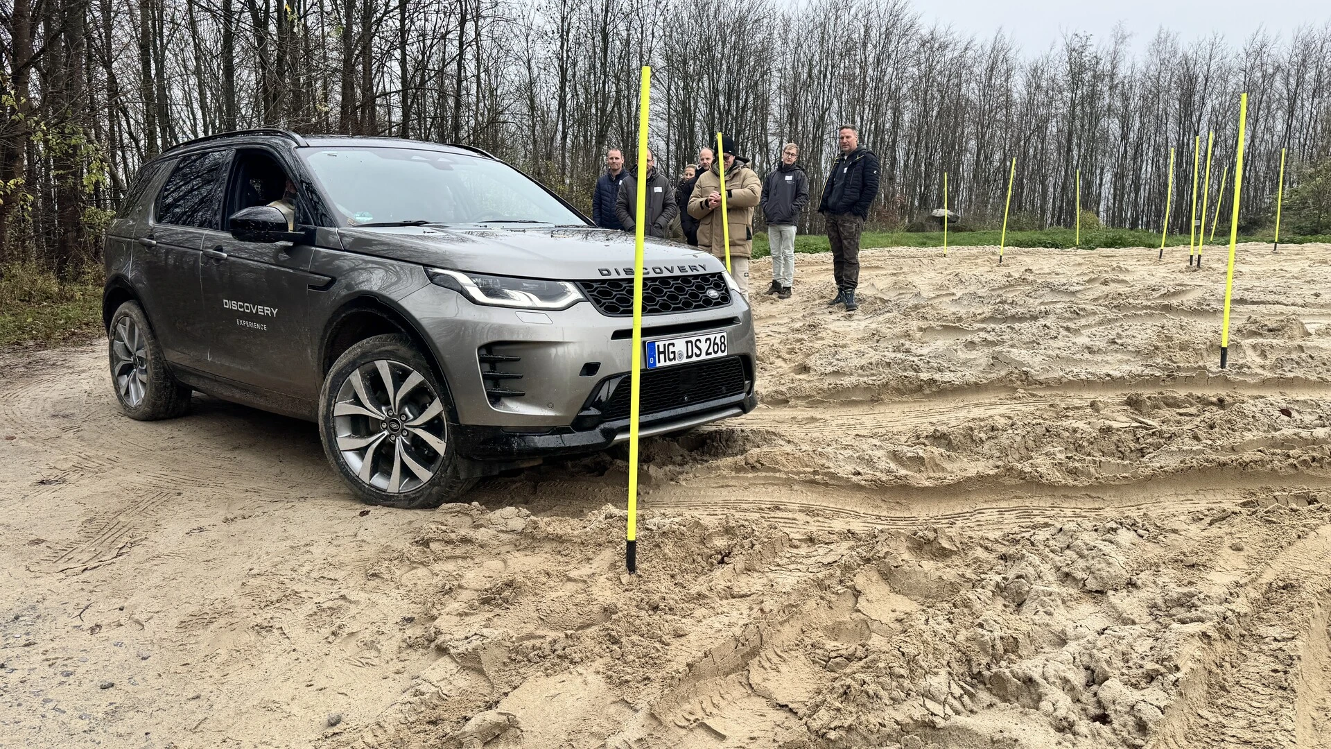 Geländewagen fährt durch einen sandigen Offroad-Parcours mit gelben Markierungsstangen. Im Hintergrund stehen mehrere Personen und beobachten das Fahrzeug