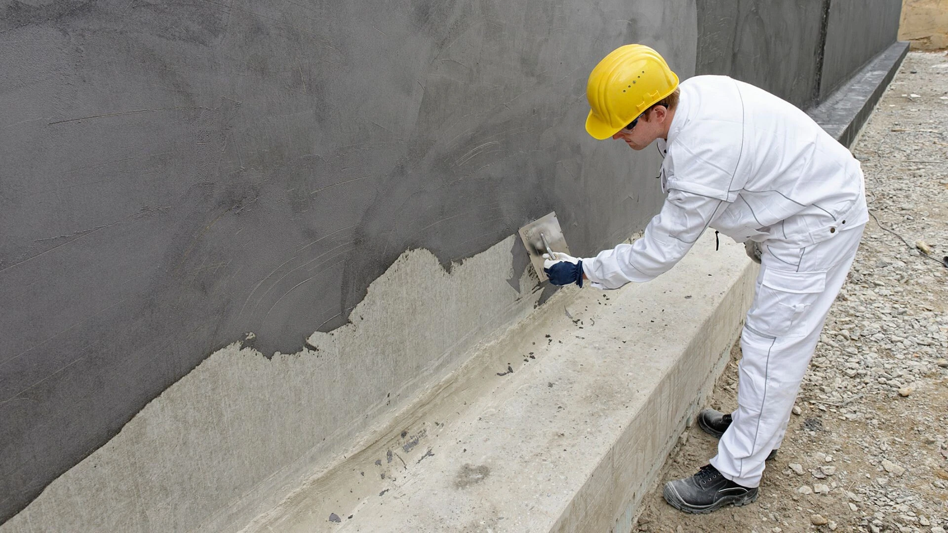 Handwerker trägt Abdichtung mit Glättkelle auf eine Betonwand im Sockelbereich eines Gebäudes