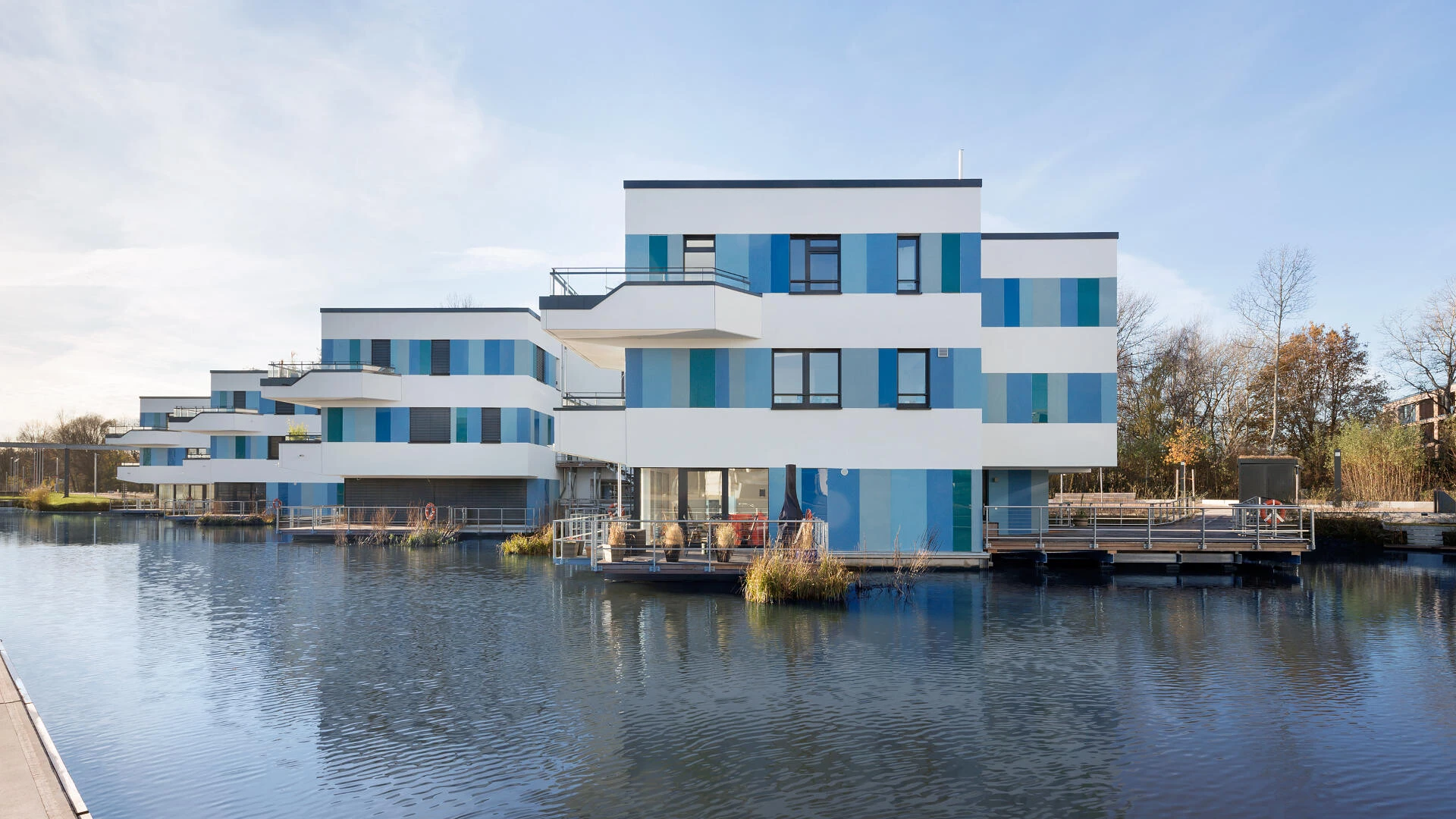 Häuserkomplex am Rand eines Sees mit einer weißen Fassade wo blaue Glasplatten drauf verklebt sind