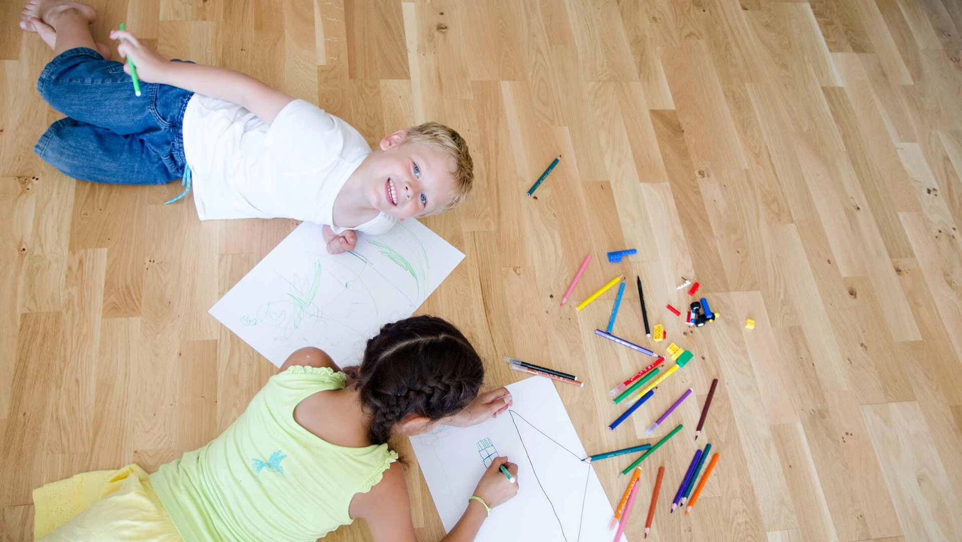 Mädchen und Junge liegen auf einem Holzfußboden nebeneinander und malen auf weißes Papier. Auf dem Boden liegen viele Buntstifte verteilt