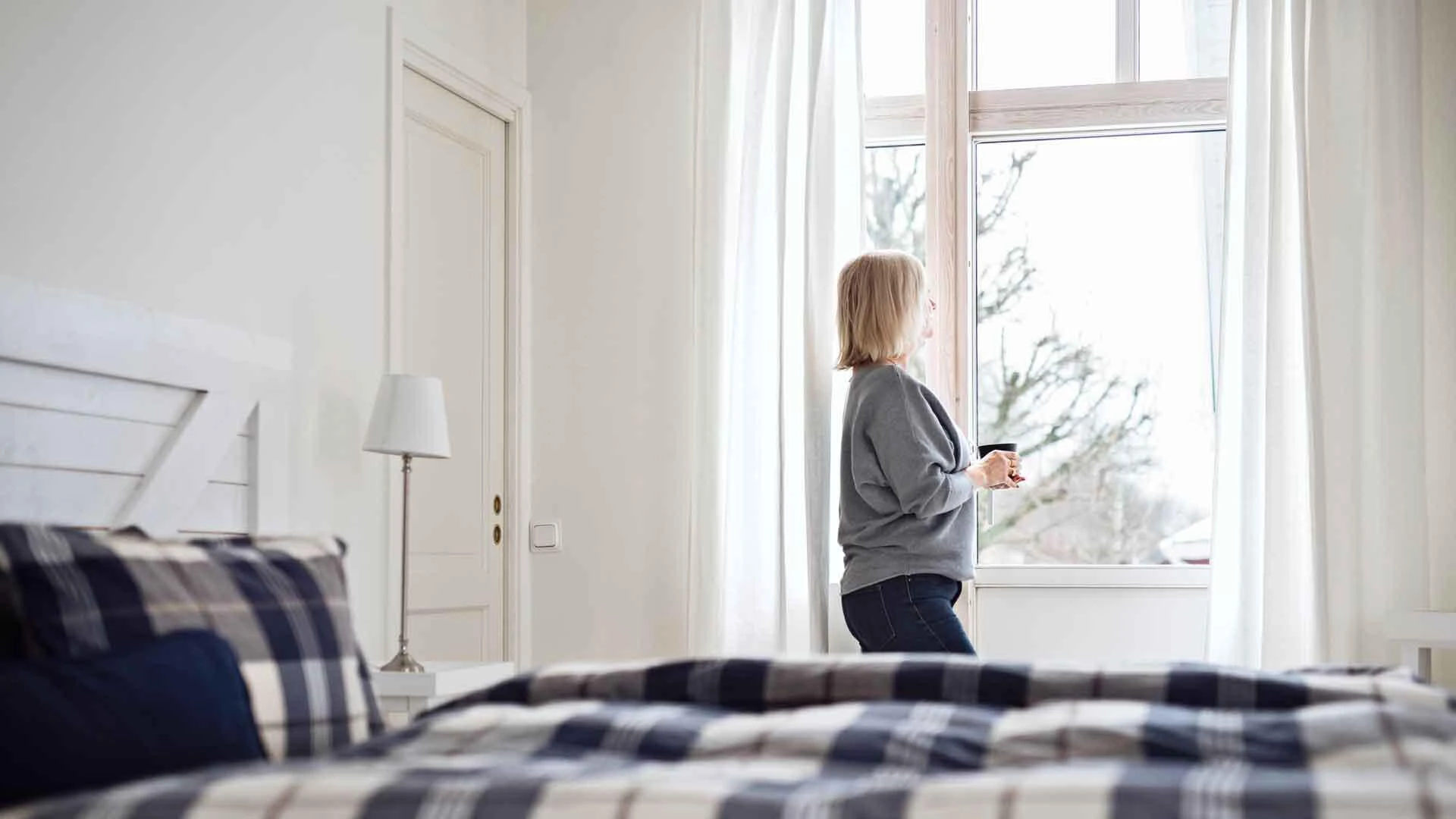 Frau steht in einem hellen Schlafzimmer am Fenster. Im Vordergrund ist blau karierte Bettwäsche auf dem Bett