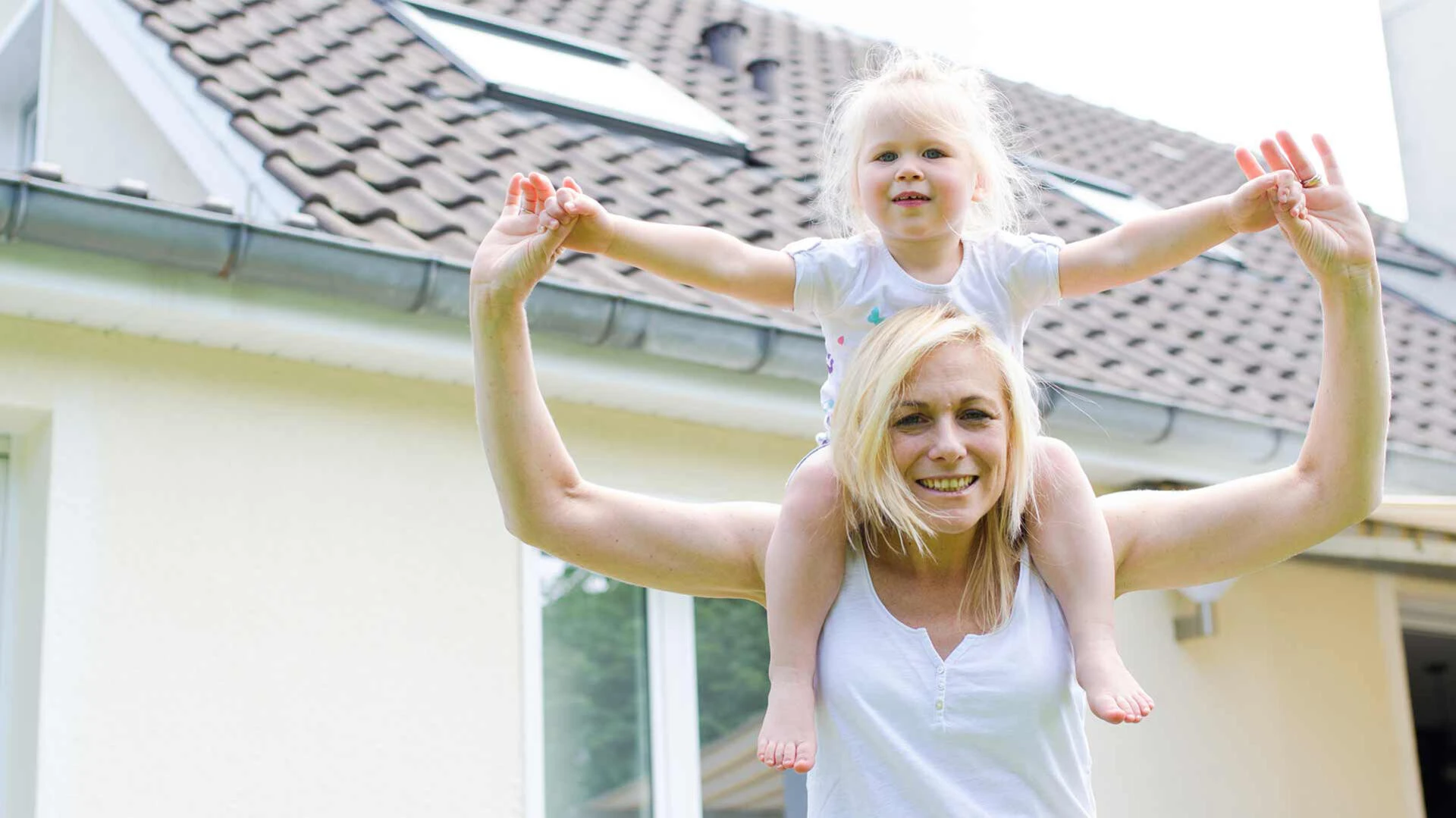 Mutter trägt ihre Tochter auf den Schultern und steht vor einem Haus mit gelber Wand