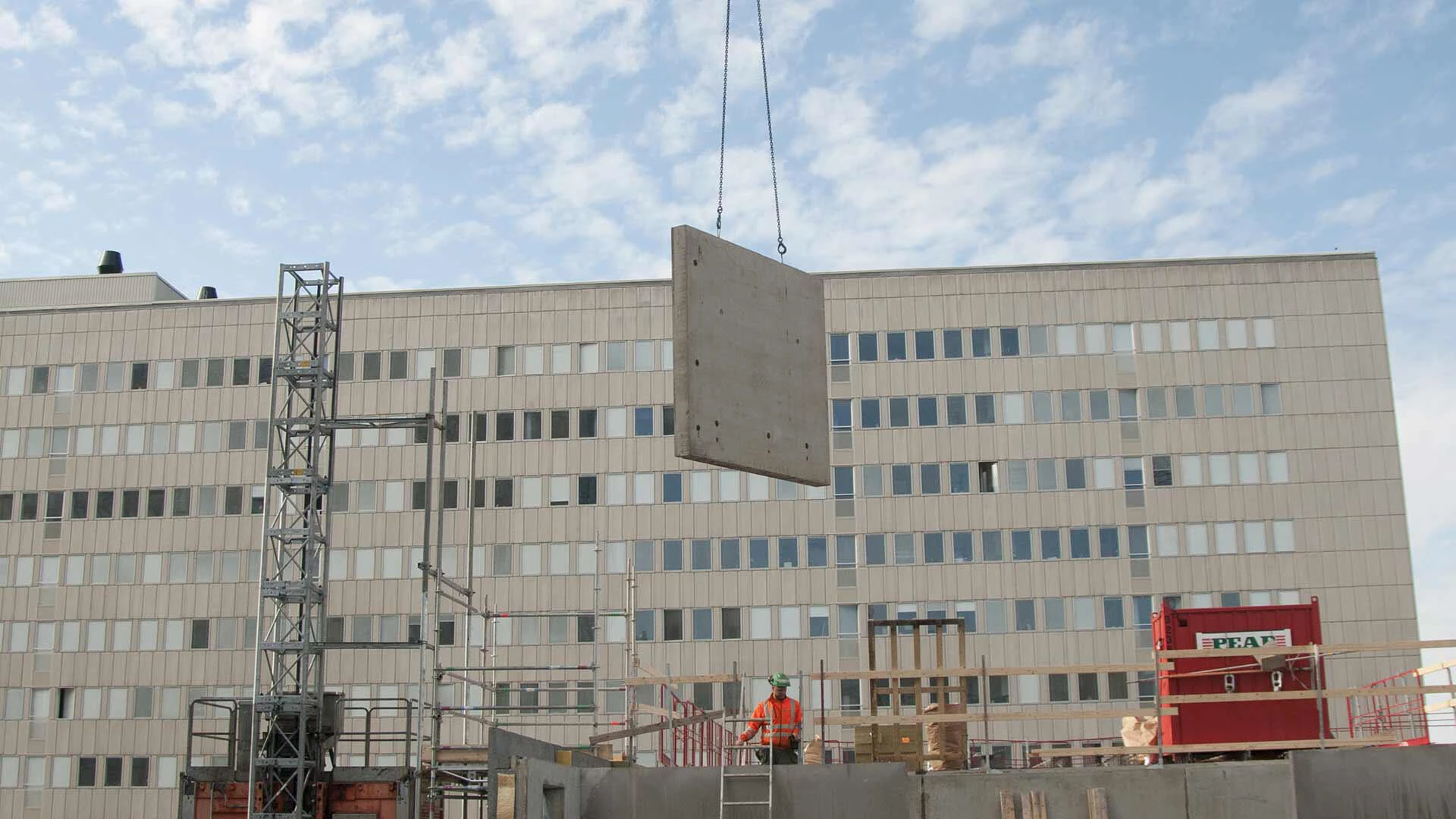 Präfabrikation hängt an einem Kran über einer Baustelle. Im Hintergrund ist ein Bürogebäude