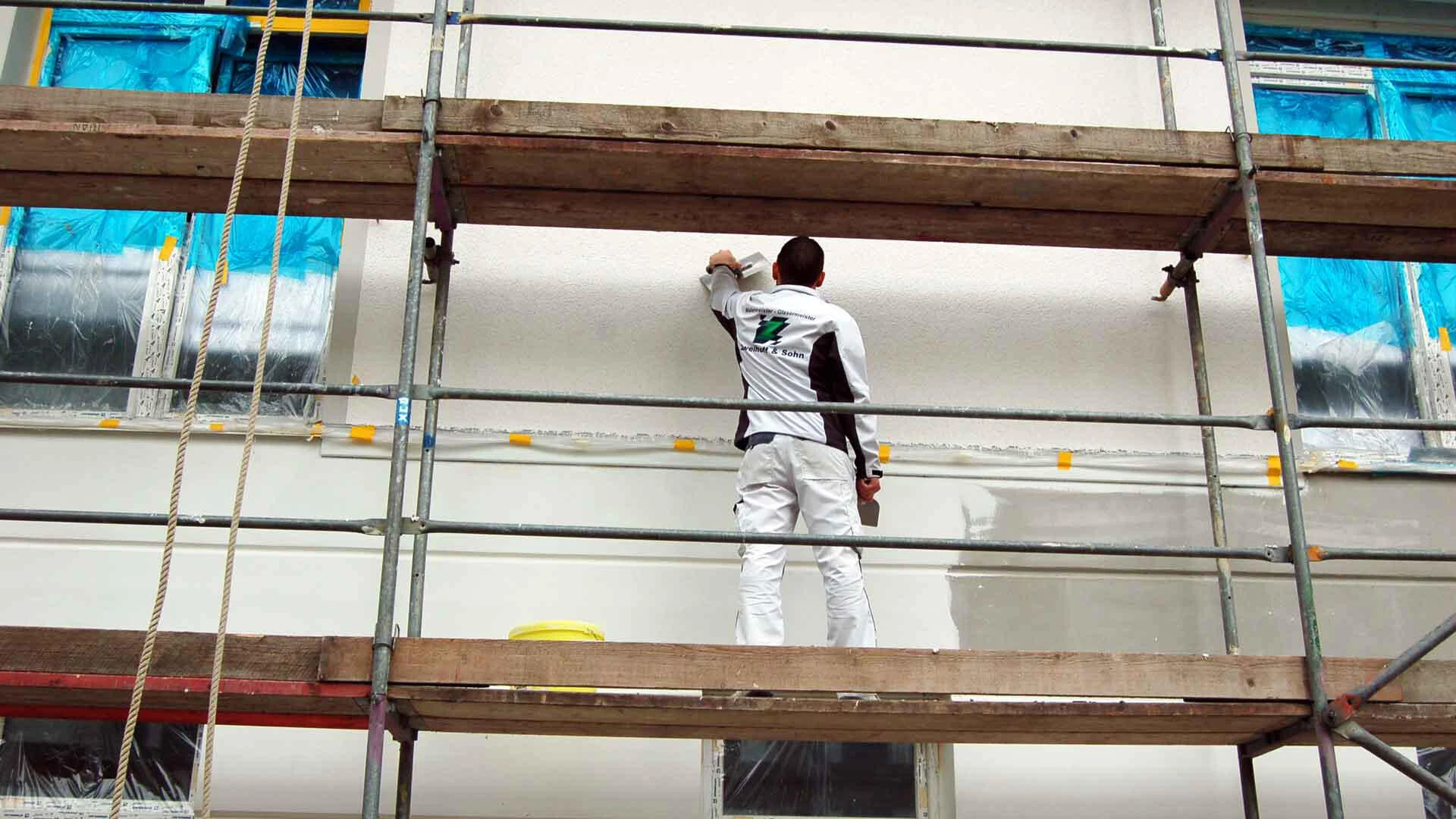Handwerker steht auf einem Gerüst an der Gebäudefassade und trägt einen weißen Putz auf