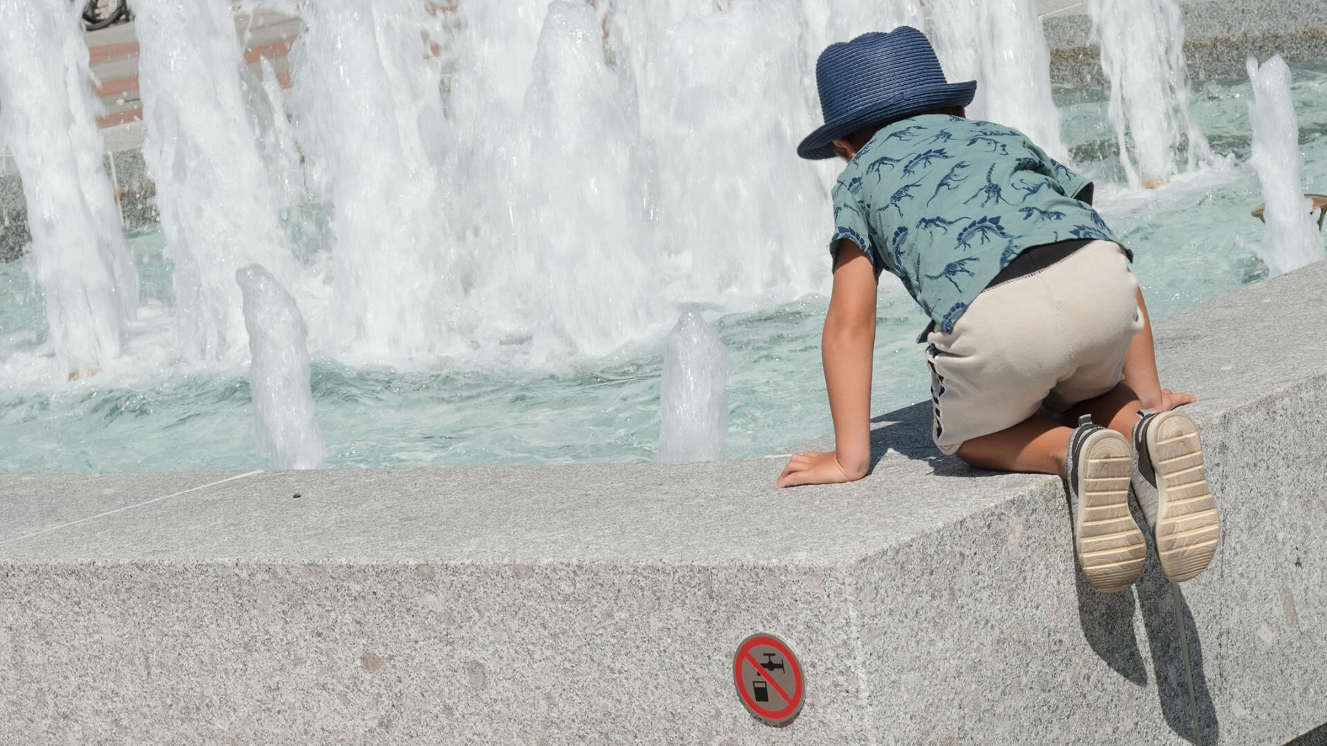 Kleines Kind mit Hut sitzt auf der Natursteinumrandung des Brunnens und guckt sich das Wasserspiel an