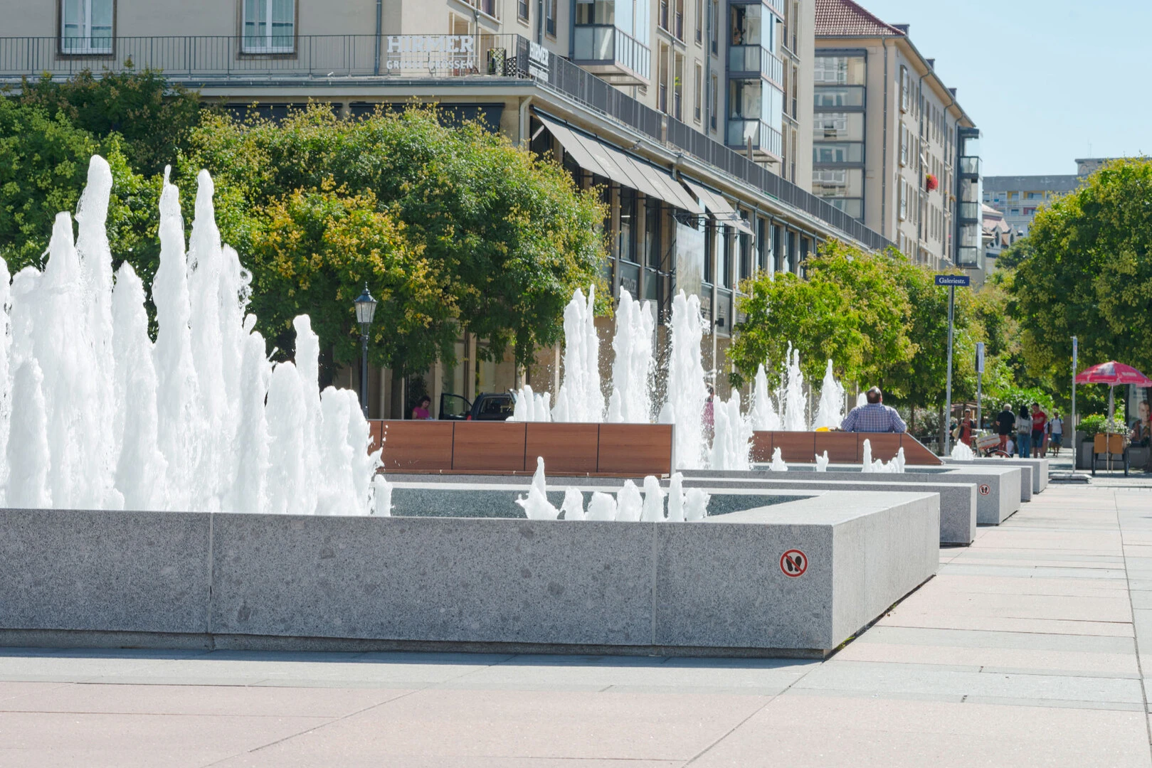Aufnahme des Brunnens vor dem Kulturpalast Dresden von weiter weg mit Blick auf die Steinbänke rund um den Brunnen