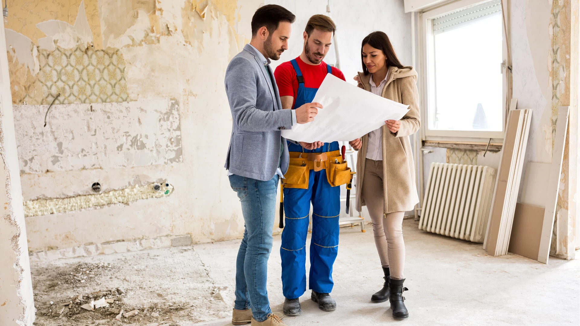 Zwei Männer und eine Frau stehen in einer Baustelle und schauen gemeinsam auf einen Plan. Ein Mann und die Frau sind schicker gekleidet, während der andere Mann Bauarbeiterkleidung trägt
