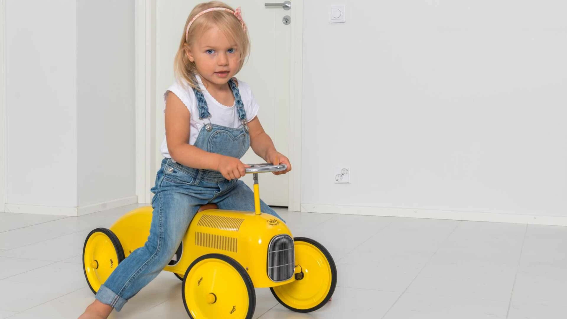 Kleines Mädchen sitzt auf einem gelben Spielauto in einem weißen Kellerraum