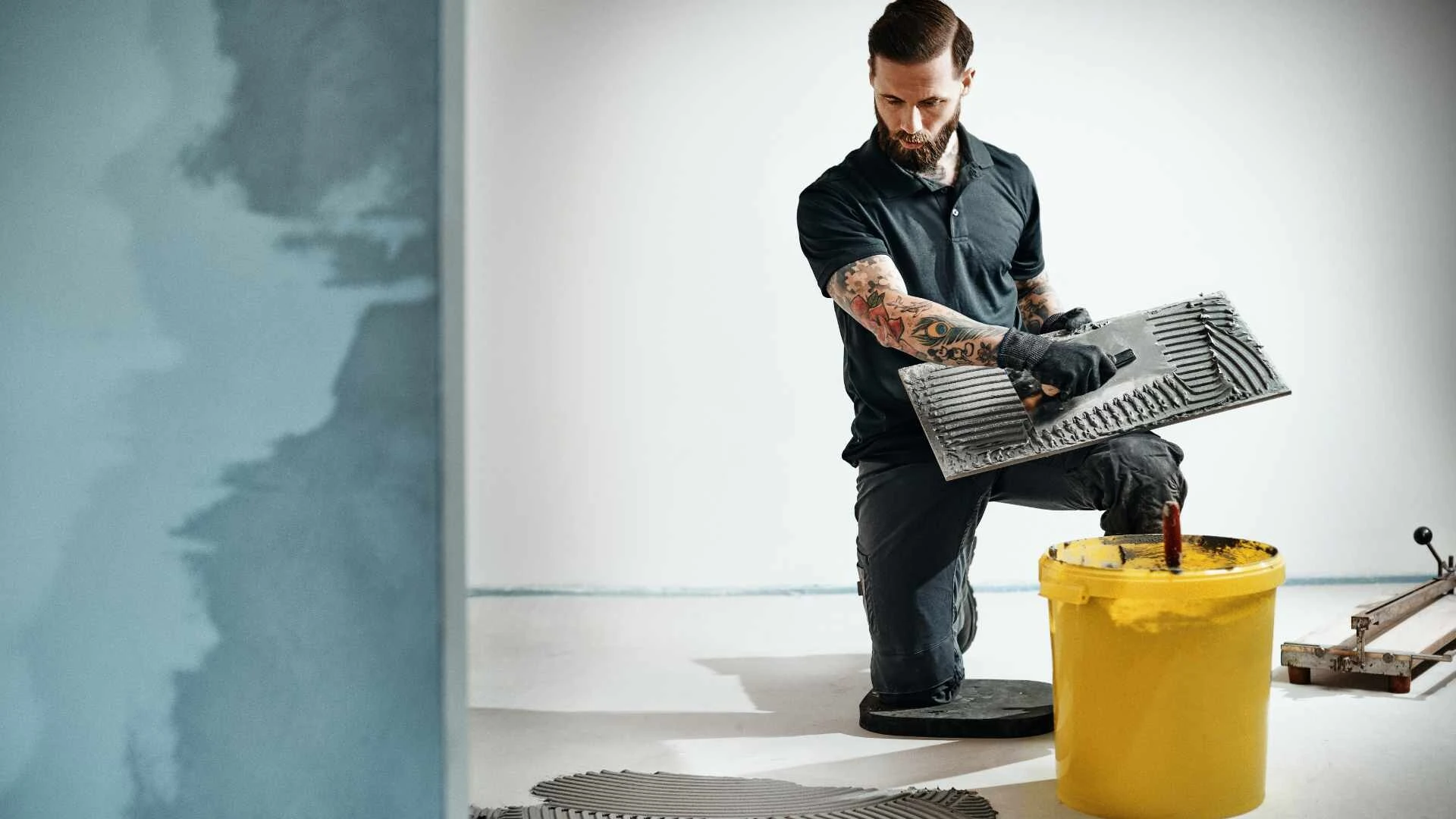 Fliesenleger kniet in einem Raum und zieht einen Kleber der BlueComfort-Produktfamilie auf die Rückseite einer Fliese für eine Badsanierung