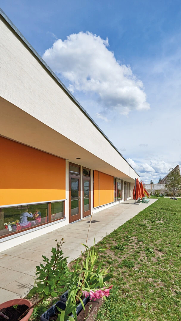 Außenfassade einer Kita mit Raffstoren vor den Fenstern und einem grünen Rasen davor