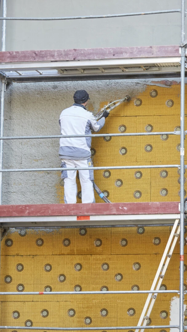 Bild, wie Handwerker auf einem Gerüst vor einer gedämmten Fassade steht und Armierungsmörtel aufspritzt