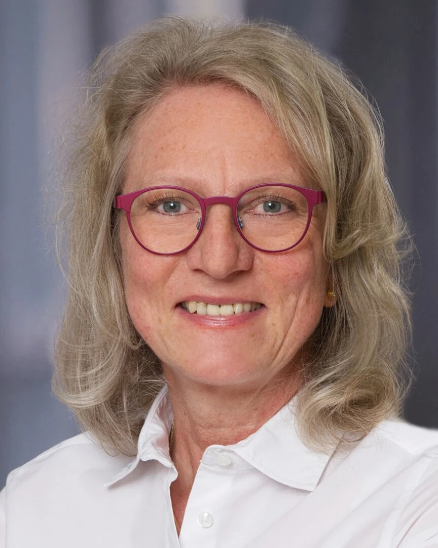 Headshot einer Frau mit blonden, schulterlangen Haaren, einer roten Brille und einer weißen Bluse