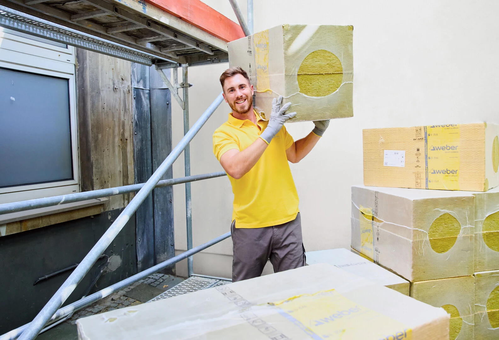 Mann in gelbem T-Shirt steht in der Mitte des Bildes und trägt auf der linken Schulter einen Pack weber.therm express Dämmplatten. Rechts im Hintergrund sind weitere Pakete der Dämmplatte zu sehen, während links eine ungedämmte Fassade mit Gerüst davor erkennbar ist.