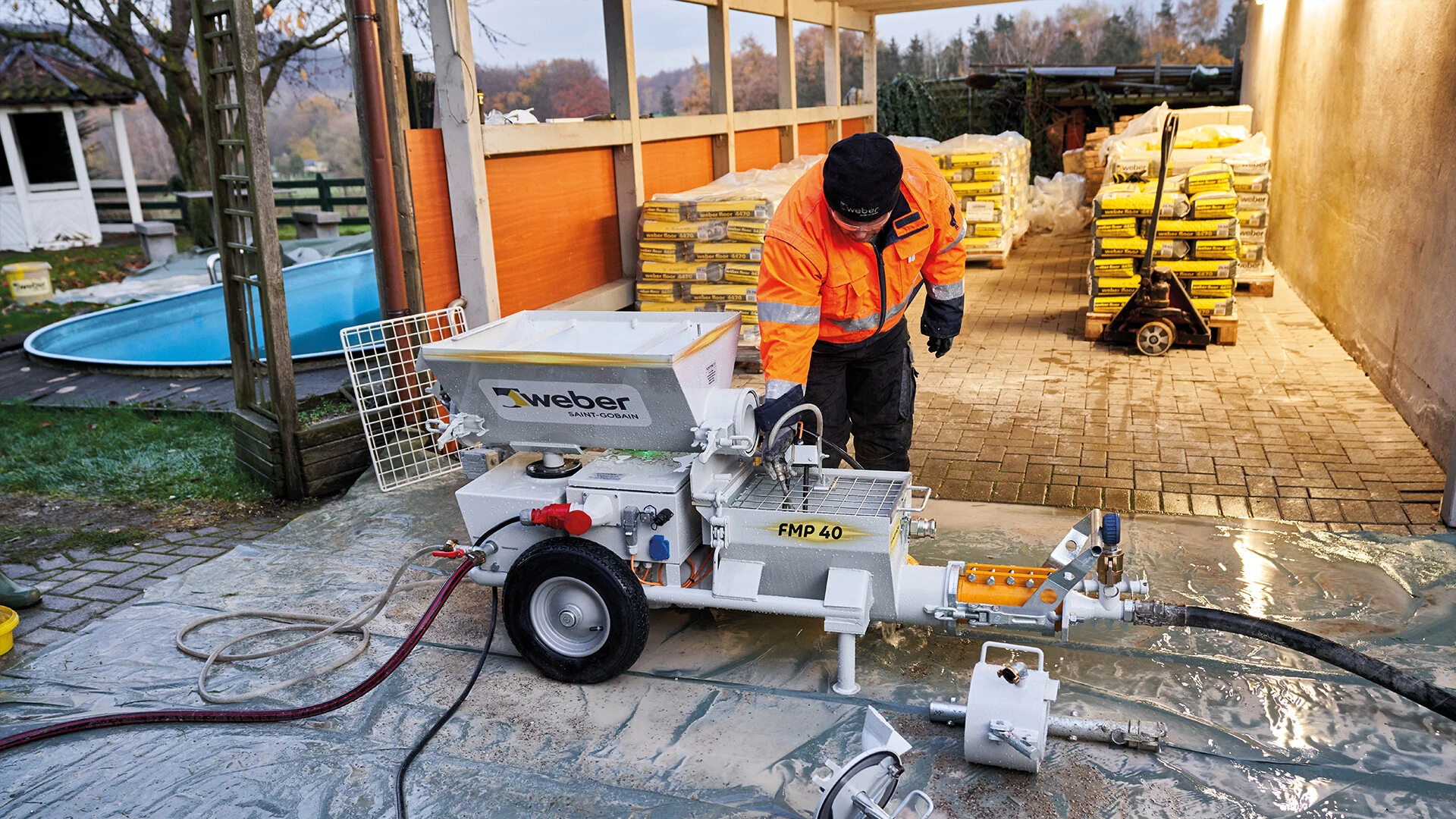 ein mann in orangener jacke steht vpr der maschinentechnik von weber, im hintergrund sieht man ein carport mit materialien