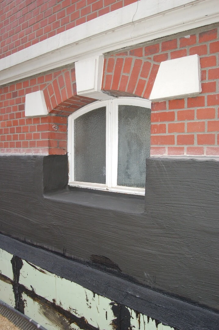 Fassadendetail eines Gebäudes mit rotem Backsteinmauerwerk und weißem Fensterrahmen. Der Bereich unterhalb des Fensters ist mit einer dunklen Abdichtungsschicht versehen, die sich deutlich vom Mauerwerk abhebt. Darunter ist eine zusätzliche Schutzschicht sichtbar, teilweise mit Bitumen beschichtet