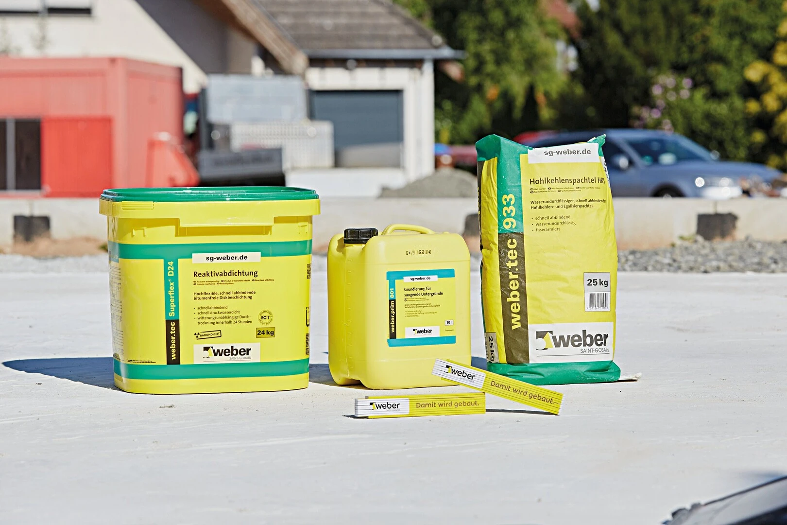 Produkte für Bauabdichtung und Sanierung von Weber auf einer Baustelle: ein gelber Eimer mit der Aufschrift ‚Reaktivabdichtung‘, ein gelber Kanister, ein grüner Sack mit der Bezeichnung ‚Hohlkehlenspachtel‘ sowie zwei Kartuschen. Im Hintergrund sind Baustellenmaterialien und ein Gebäude sichtbar.