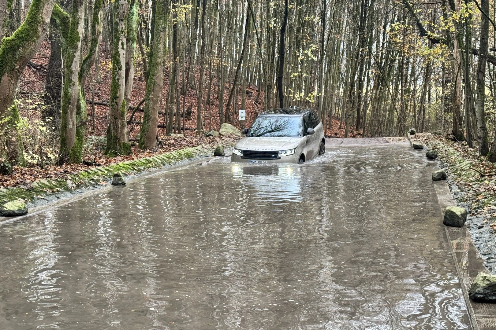 Geländewagen fährt durch eine überflutete Waldstraße, umgeben von Bäumen mit Herbstlaub. Das Wasser bedeckt fast die gesamte Fahrbahn, und die Fahrzeugfront ist mit eingeschaltetem Licht sichtbar