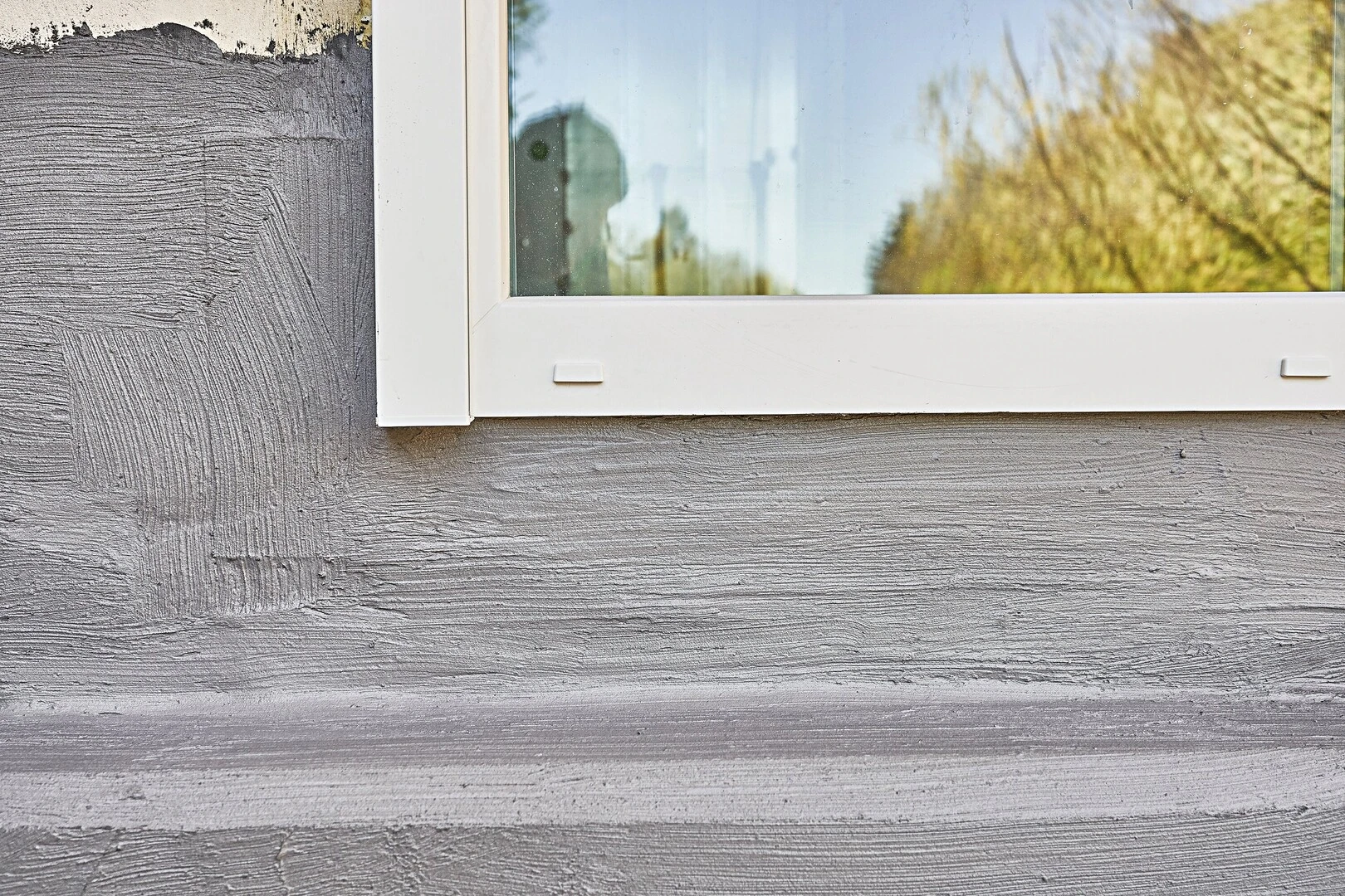 Detailaufnahme einer strukturierten Putzoberfläche unterhalb eines weißen Fensterrahmens. Die Oberfläche zeigt feine horizontale Rillen, die durch Bürstenbearbeitung entstanden sind. Im Fenster spiegelt sich die Umgebung mit Bäumen und Himmel.