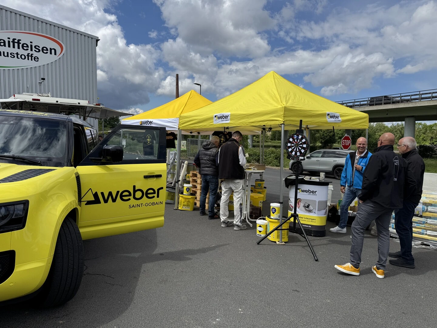 Outdoor-Event mit gelben Saint Gobain Weber Pavillons auf einem Parkplatz. Unter den Pavillons stehen mehrere Eimer mit Bauprodukten, ein Tisch mit Werbematerialien und eine Dartscheibe auf einem Stativ. Links im Vordergrund ist ein gelbes Fahrzeug mit Weber-Logo und geöffneter Tür zu sehen. Im Hintergrund sind ein Raiffeisen-Baustoffe-Gebäude, eine Straße mit Brücke und ein teilweise bewölkter Himmel.
