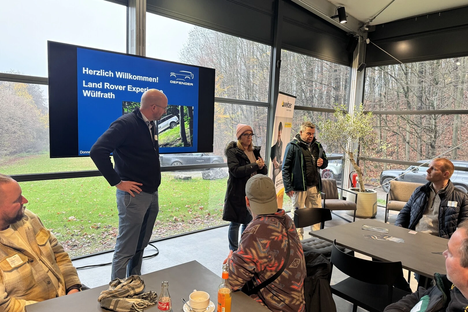 Innenaufnahme eines Veranstaltungsraums mit großen Fenstern, Blick ins Grüne. Im Vordergrund ein Tisch mit Getränken und Tassen, im Hintergrund ein Bildschirm mit der Aufschrift ‚Herzlich Willkommen! Land Rover Experience Wülfrath‘. Mehrere Personen stehen und unterhalten sich.