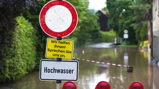 Bild von einer Straße, die wegen Hochwasser gesperrt ist. Im Vordergrund sieht man ein Stoppschild mit dem Text Hochwasser und Wir helfen Ihnen! Sprechen Sie uns an darauf.