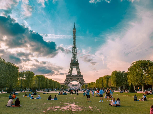 Der Eifelturm in Paris vor blauem Himmel mit ein paar weißen Wolken und davor Grüppchen von Menschen verteilt auf der grünen Wiese