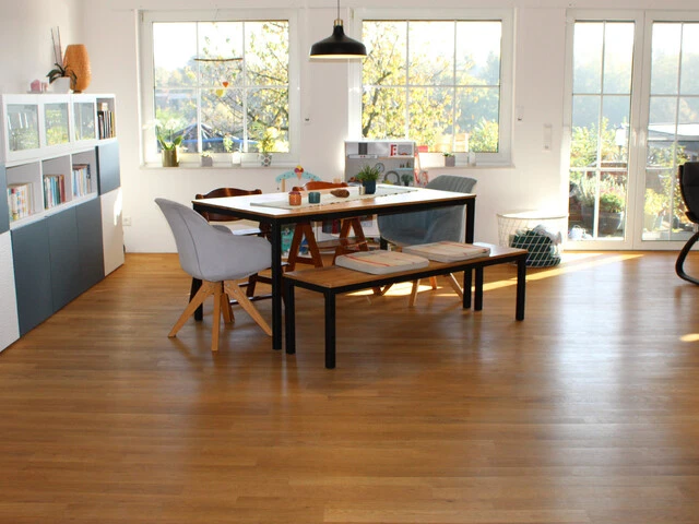 Aufnahme in ein Wohnzimmer mit einem Esstisch in der Mitte, einem Sideboard auf der linken Seite und einem Stuhl auf der rechten Seite. Beim Boden handelt es sich um einen braunen Holzboden und im Hintergrund sind große Fenster zu erkennen.