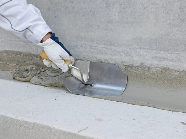 Nahaufnahme einer Hand, die mit einer Kelle eine mineralische Hohlkehle im Übergangsbereich zwischen einem Kellerboden und der -wand zieht.
