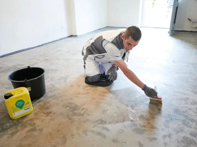 Handwerker kniet auf einem mineralischen Kellerboden und trägt mit einem Pinsel die Grundierung auf