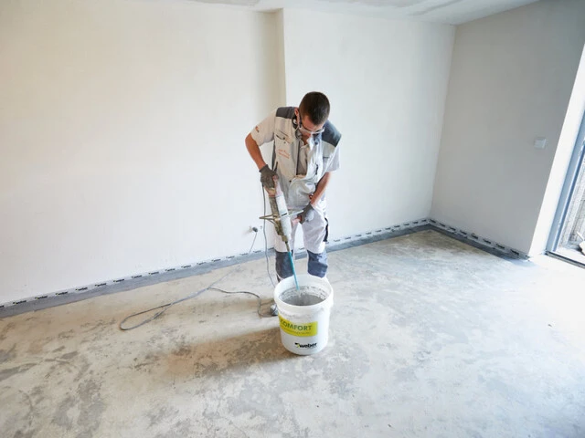 Handwerker steht in einem Kellerraum und mischt mit einem Rührgerät einen Dichtspachtel in einem Eimer an
