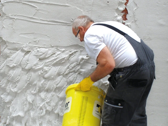 Handwerker steht vor einer Kellerwand und trägt aus einem Eimer weißen Sanierputz auf die Wand auf