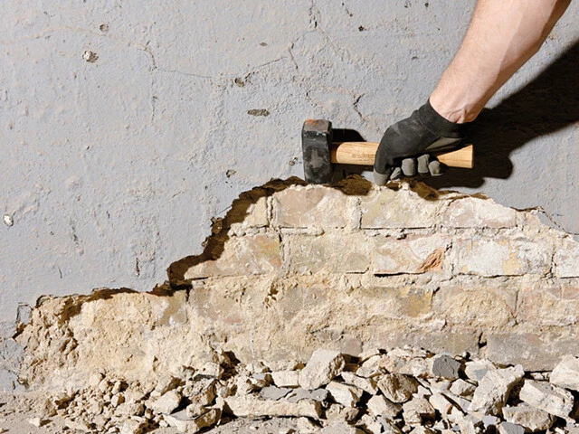 Aufnahme einer Hand, die mit einem Hammer Altputz von einer Kellerwand abschlägt